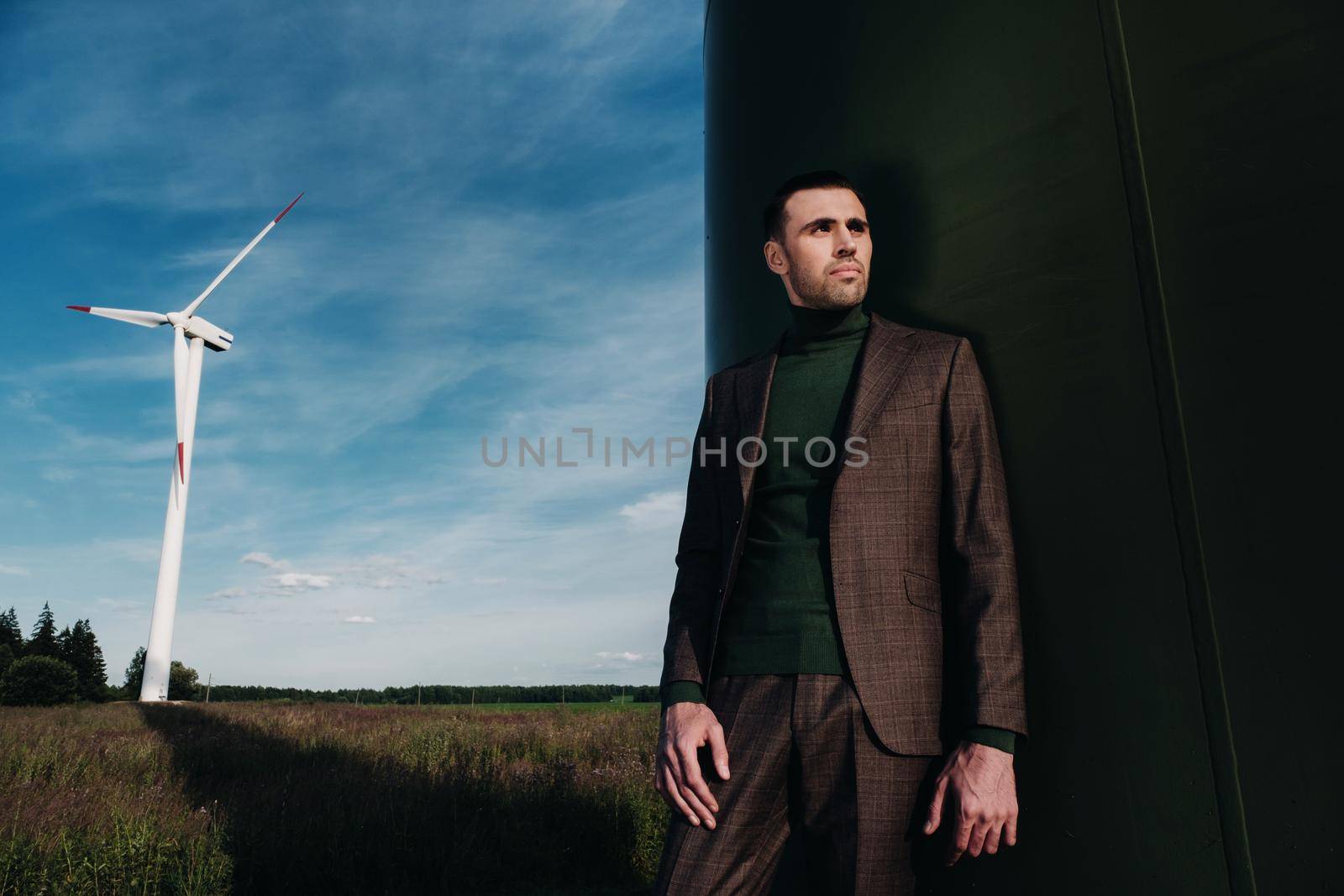 A man in a business suit with a green Golf shirt stands next to a windmill against the background of the field and the blue sky.Businessman near the windmills.Modern concept of the future. by Lobachad