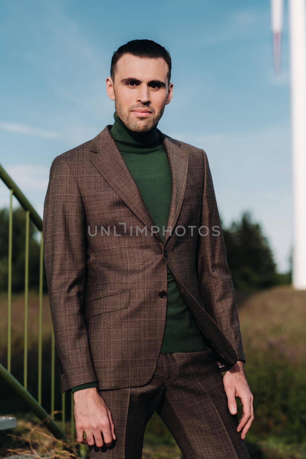 A man in a business suit with a green Golf shirt stands next to a windmill against the background of the field and the blue sky.Businessman near the windmills.Modern concept of the future. by Lobachad