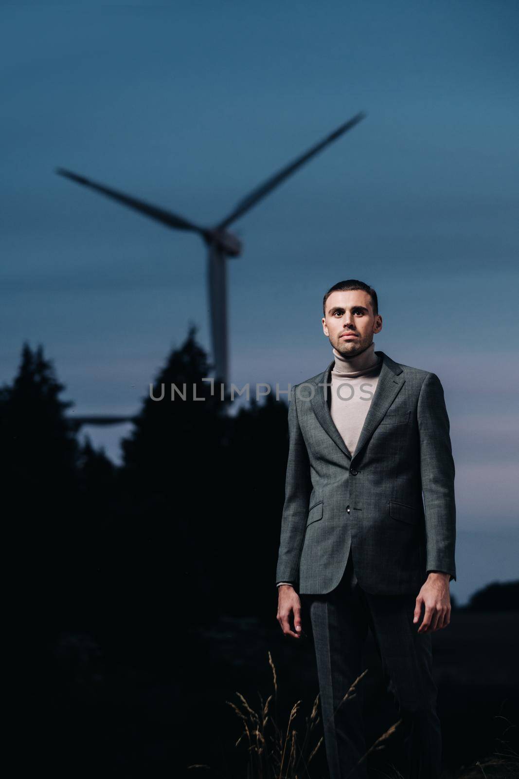 A man in a grey business suit stands next to a windmill after sunset .Businessman near windmills at night.Modern concept of the future. by Lobachad