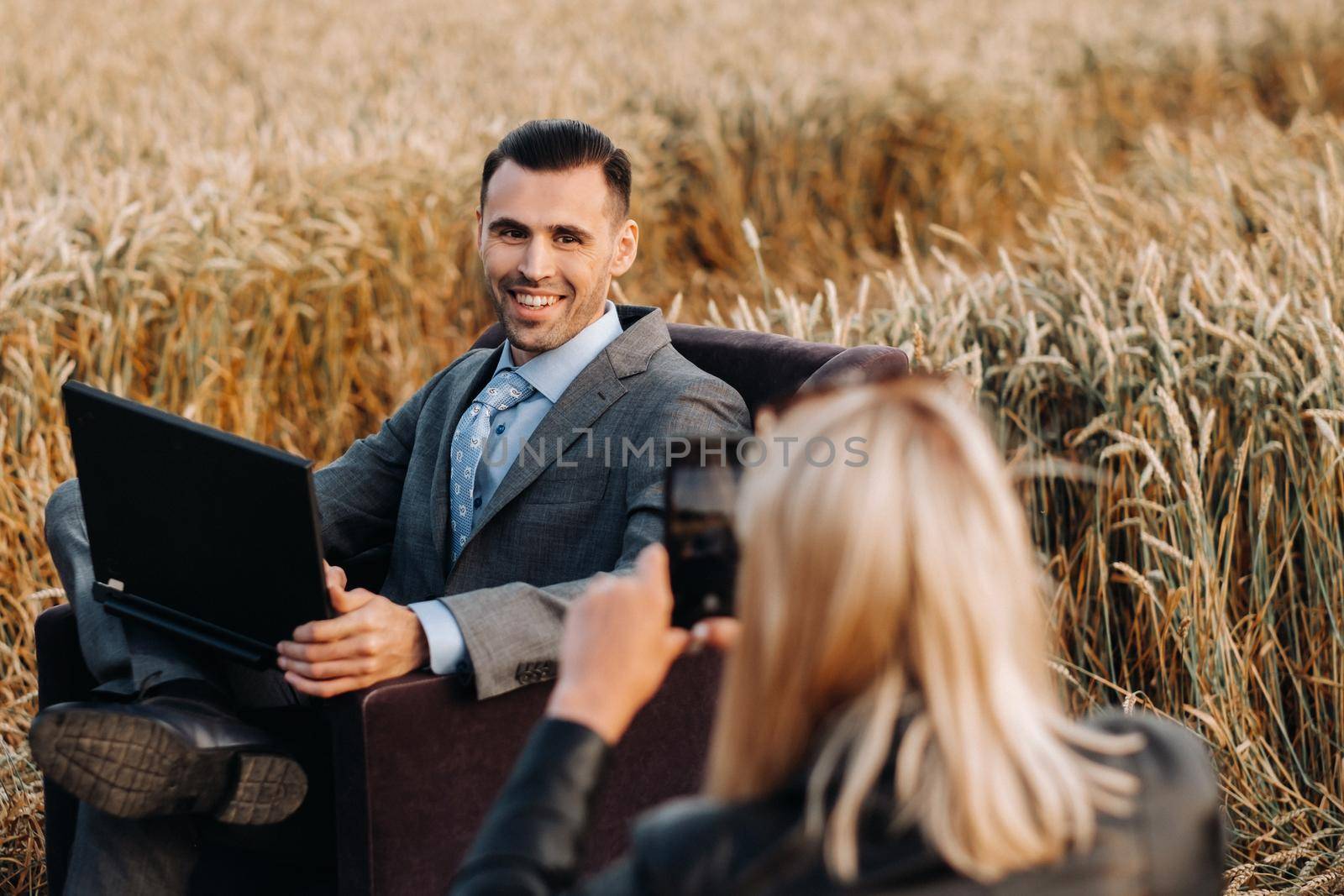 Portrait of a businessman in a suit with a laptop sitting in a chair in a wheat field and photographed by an unrecognizable girl. by Lobachad