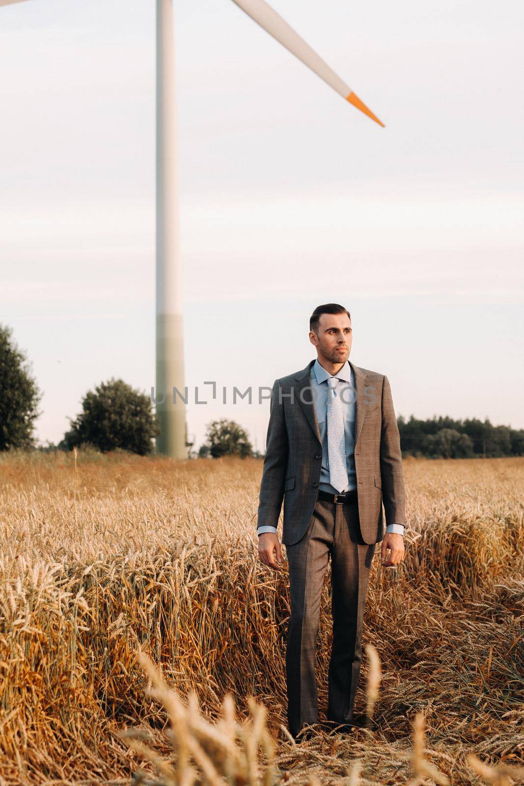 Portrait of a businessman in a gray suit on a wheat field against the background of a windmill and the evening sky.