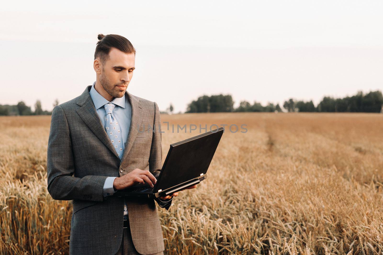 Portrait of a businessman in a suit holding a laptop in a field of wheat against the background of a windmill and the evening sky.