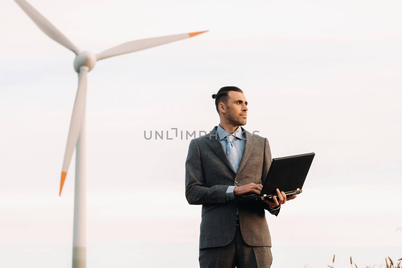 Portrait of a businessman in a suit holding a laptop in a field of wheat against the background of a windmill and the evening sky.