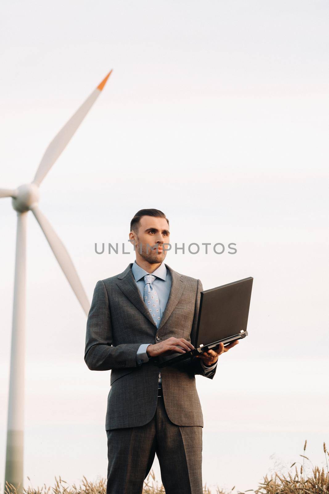 Portrait of a businessman in a suit holding a laptop in a field of wheat against the background of a windmill and the evening sky by Lobachad