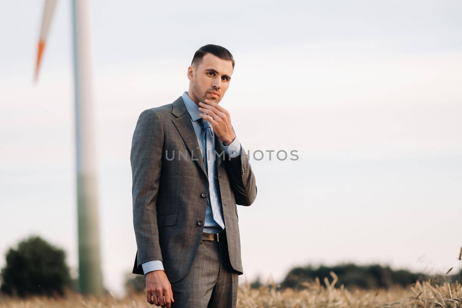 Portrait of a businessman in a gray suit on a wheat field against the background of a windmill and the evening sky.