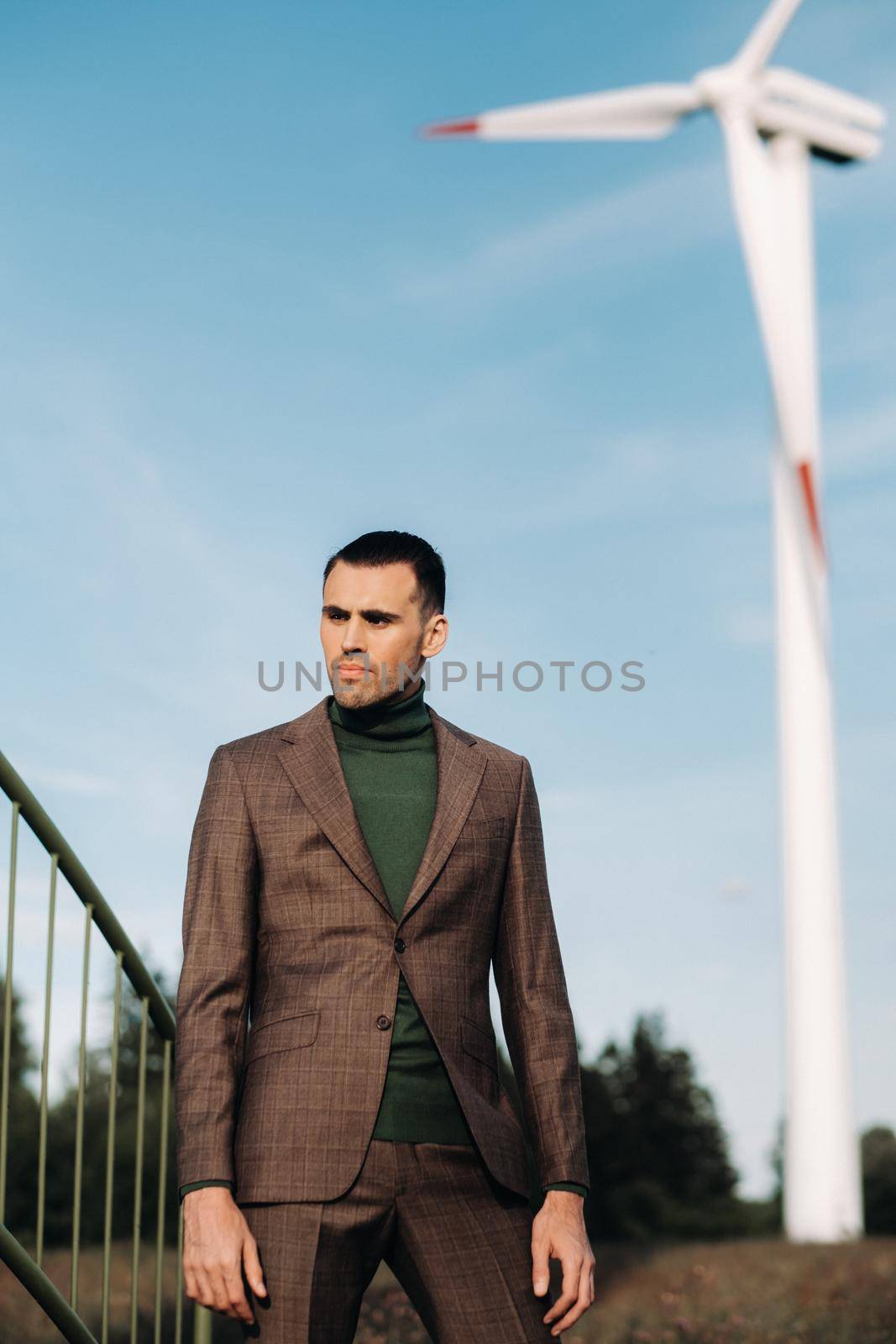 A man in a business suit with a green Golf shirt stands next to a windmill against the background of the field and the blue sky.Businessman near the windmills.Modern concept of the future