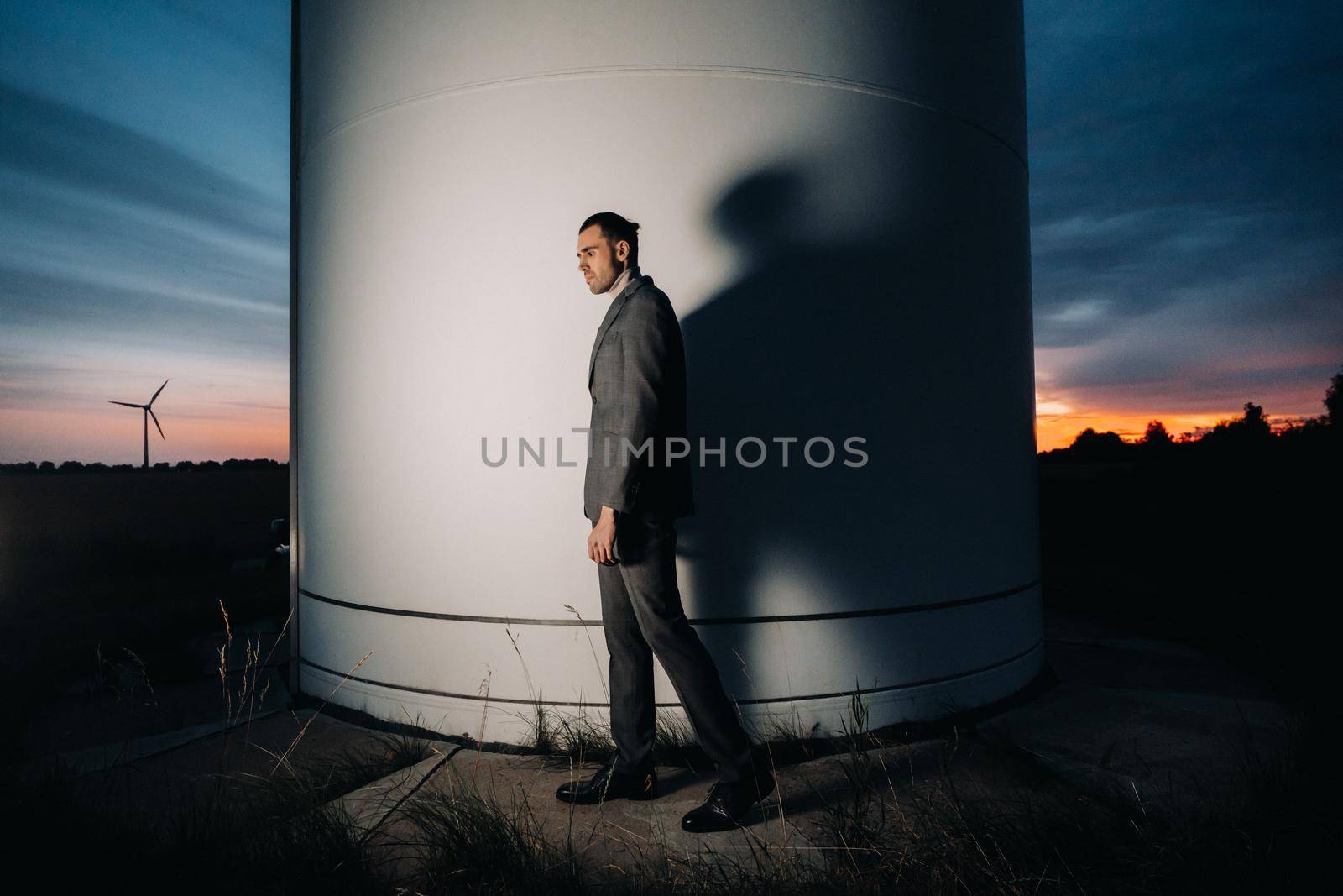 A man in a grey business suit stands next to a windmill after sunset .Businessman near windmills at night.Modern concept of the future. by Lobachad