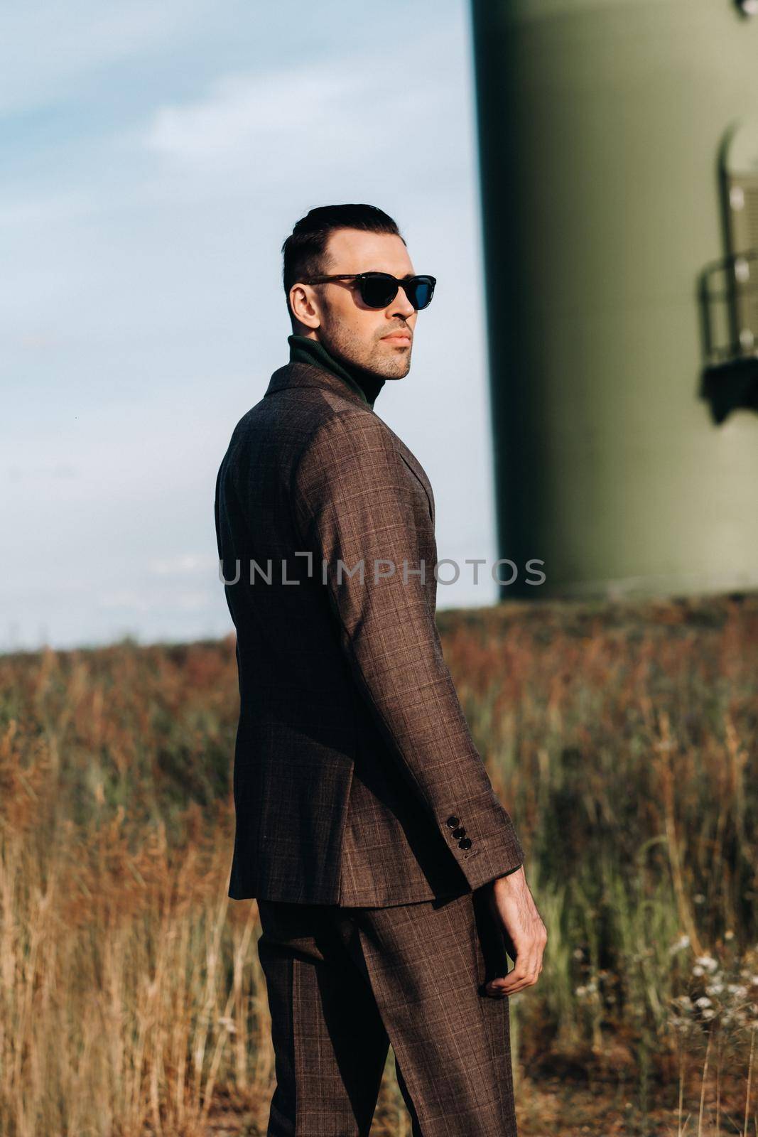 A man in a business suit stands next to a windmill against a field and blue sky.Businessman near the windmills.Modern concept of the future. by Lobachad