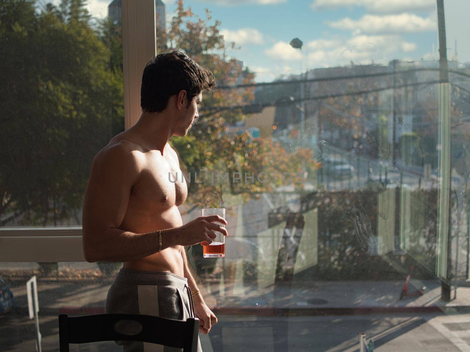 Sexy handsome young man standing shirtless,drinking a glass of soda or tea next to window