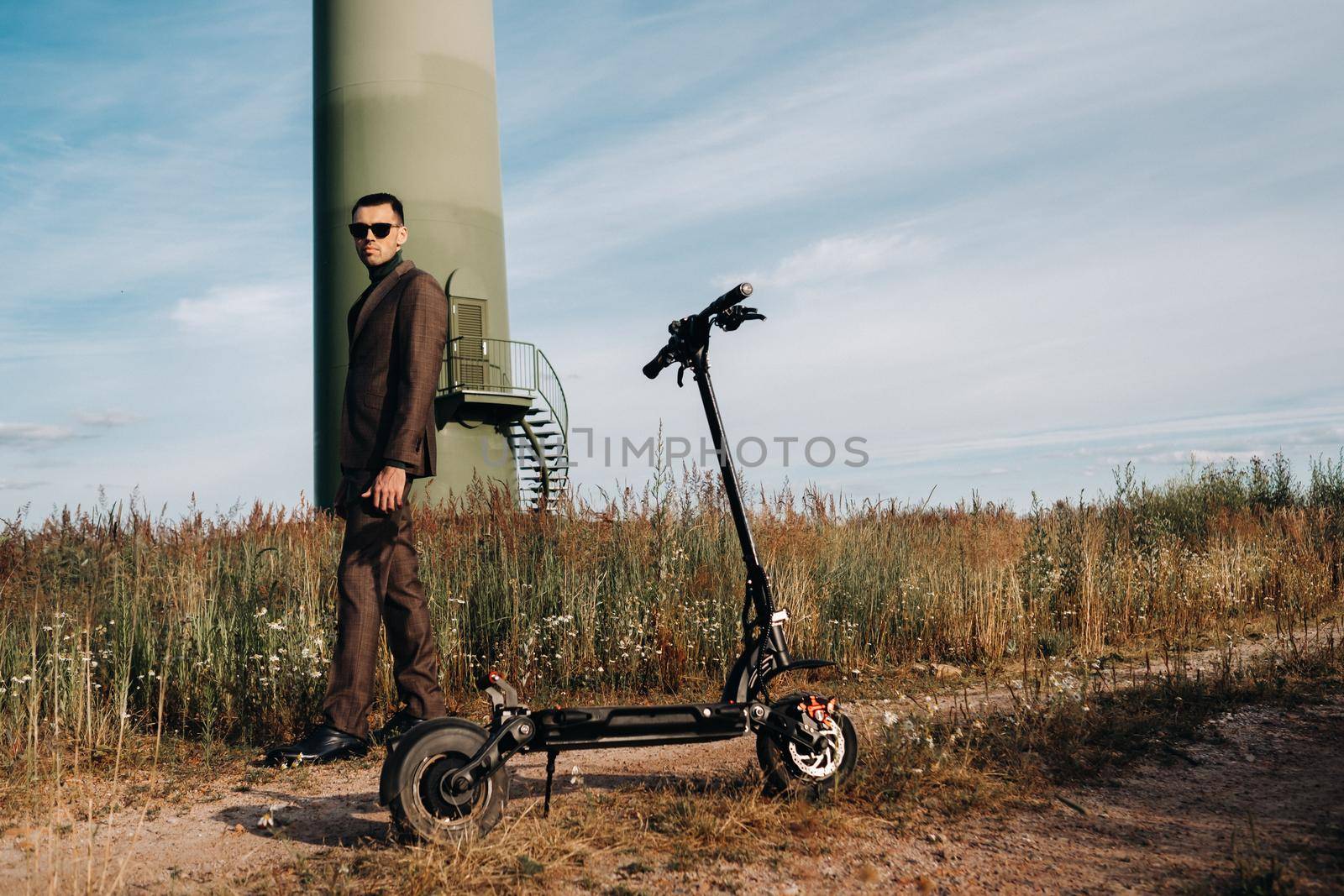 A man in a business suit and green Golf shirt stands next to an electric scooter against a field and blue sky.Businessman near the scooter.Modern concept of the future