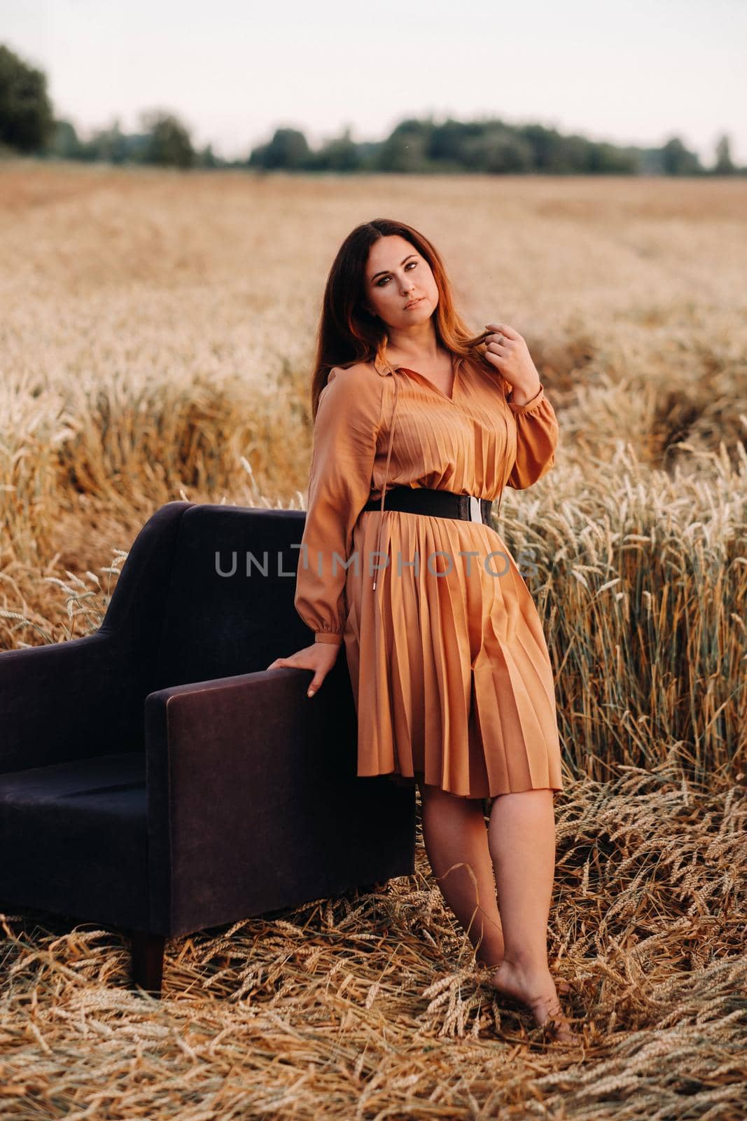 A girl in a orange dress stands next to a chair in a wheat field in the evening by Lobachad