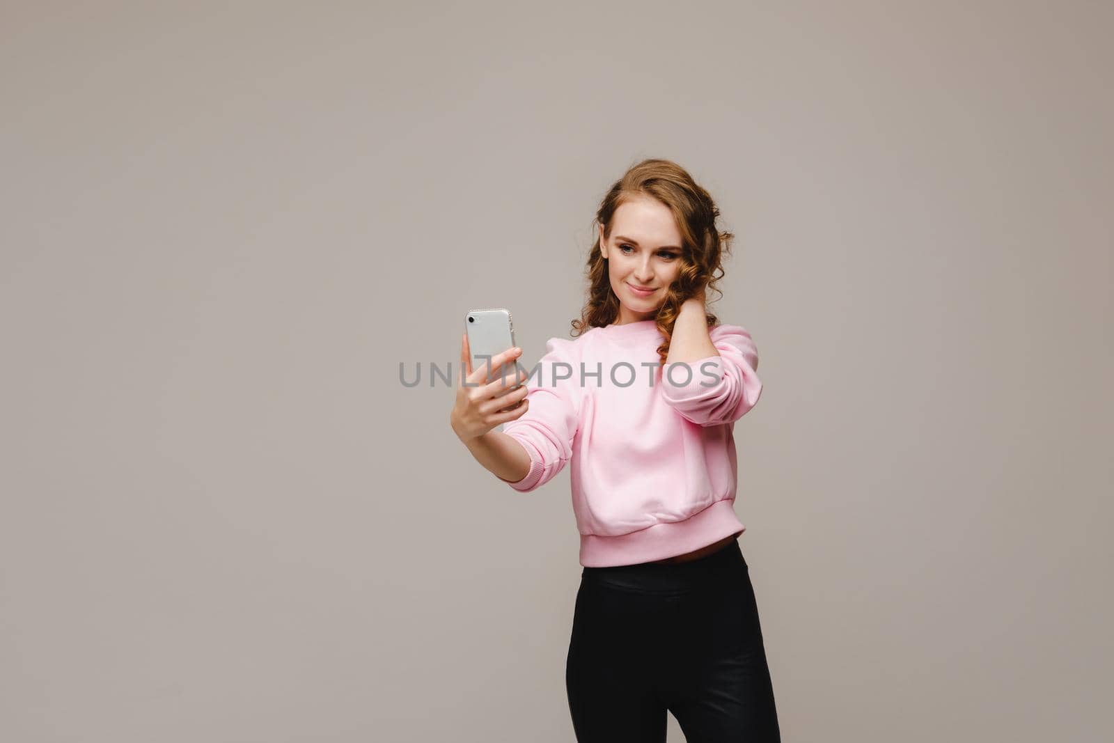 A smiling happy girl in a pink blouse takes a selfie on a smartphone on a gray background