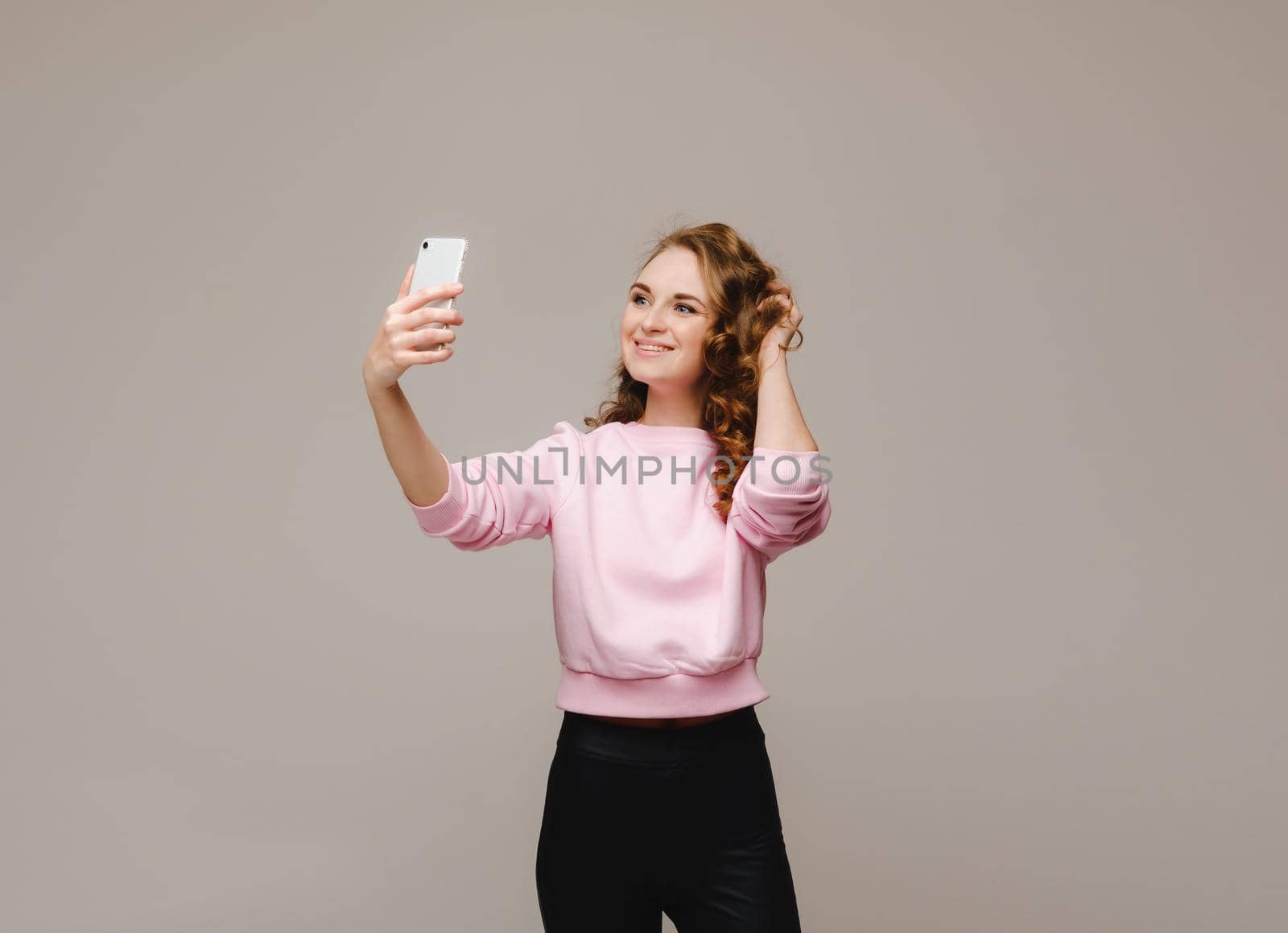 A smiling happy girl in a pink blouse takes a selfie on a smartphone on a gray background