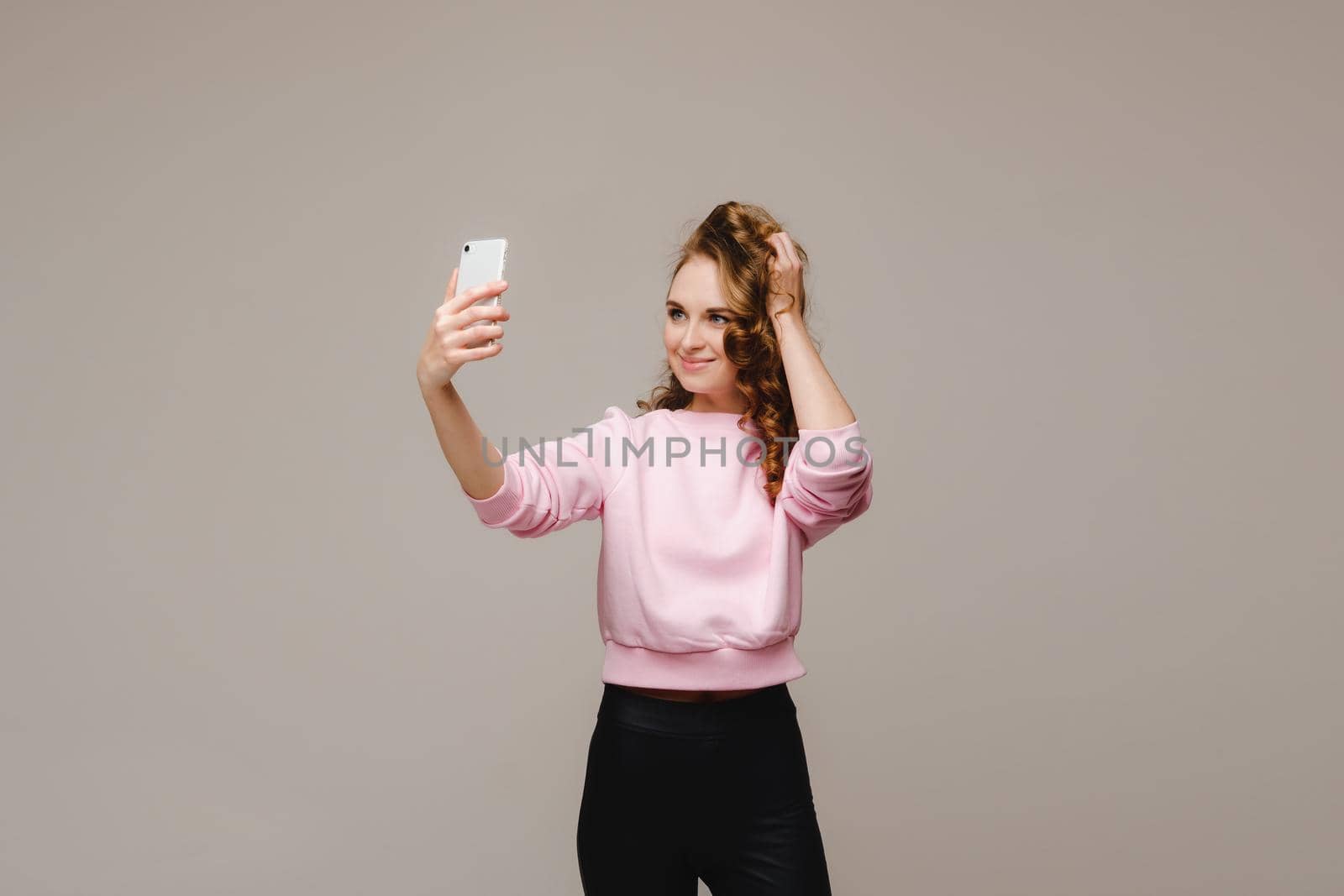 A smiling happy girl in a pink blouse takes a selfie on a smartphone on a gray background