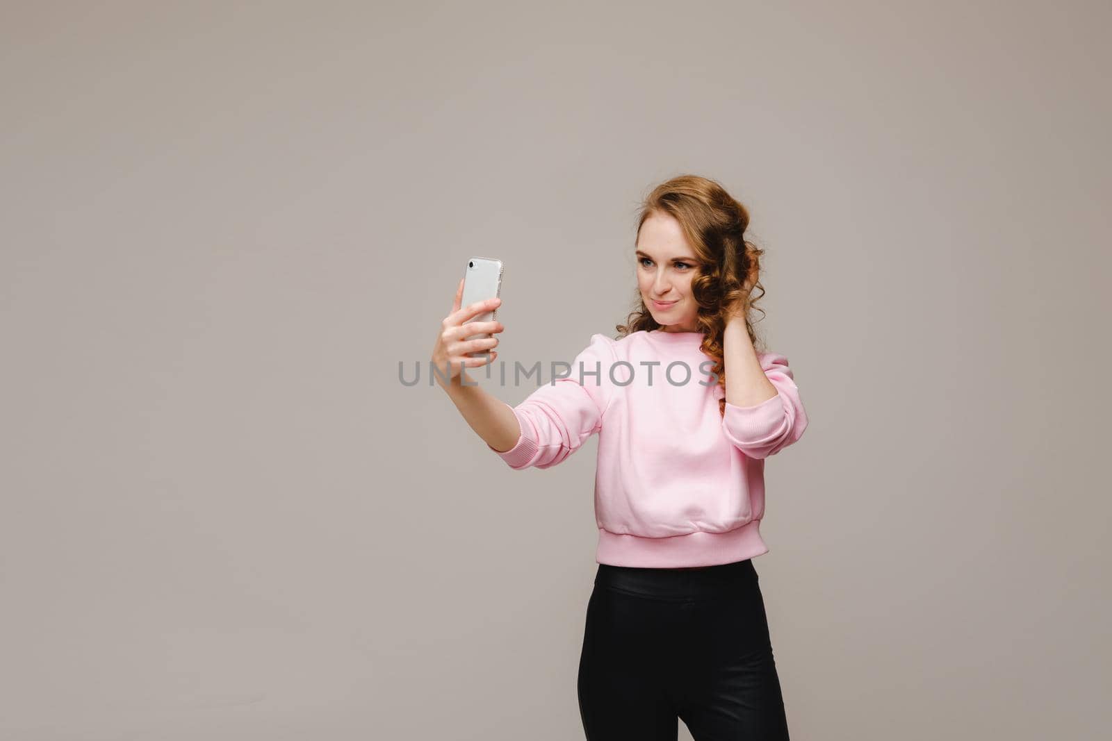 A smiling happy girl in a pink blouse takes a selfie on a smartphone on a gray background