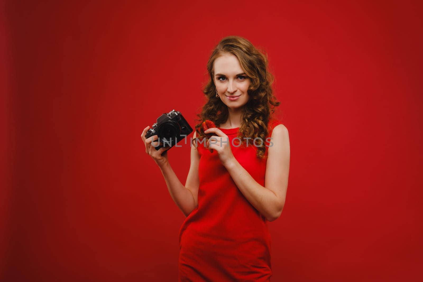 a smiling young woman with wavy hair holds a strawberry and photographs it, holding a delicious fresh strawberry on a bright red background.