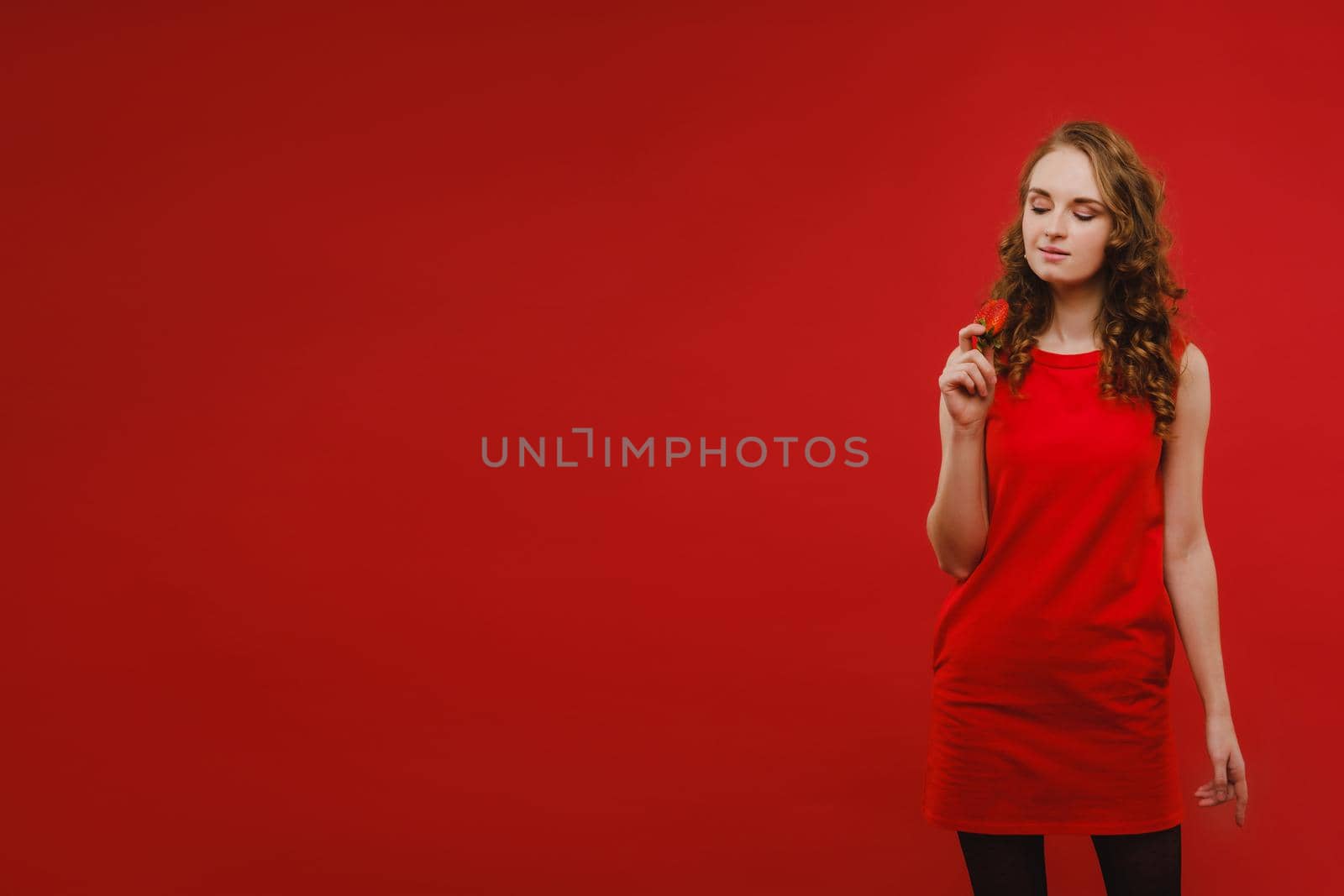 A beautiful girl in a red dress on a red background holds a strawberry in her hands and smiles.