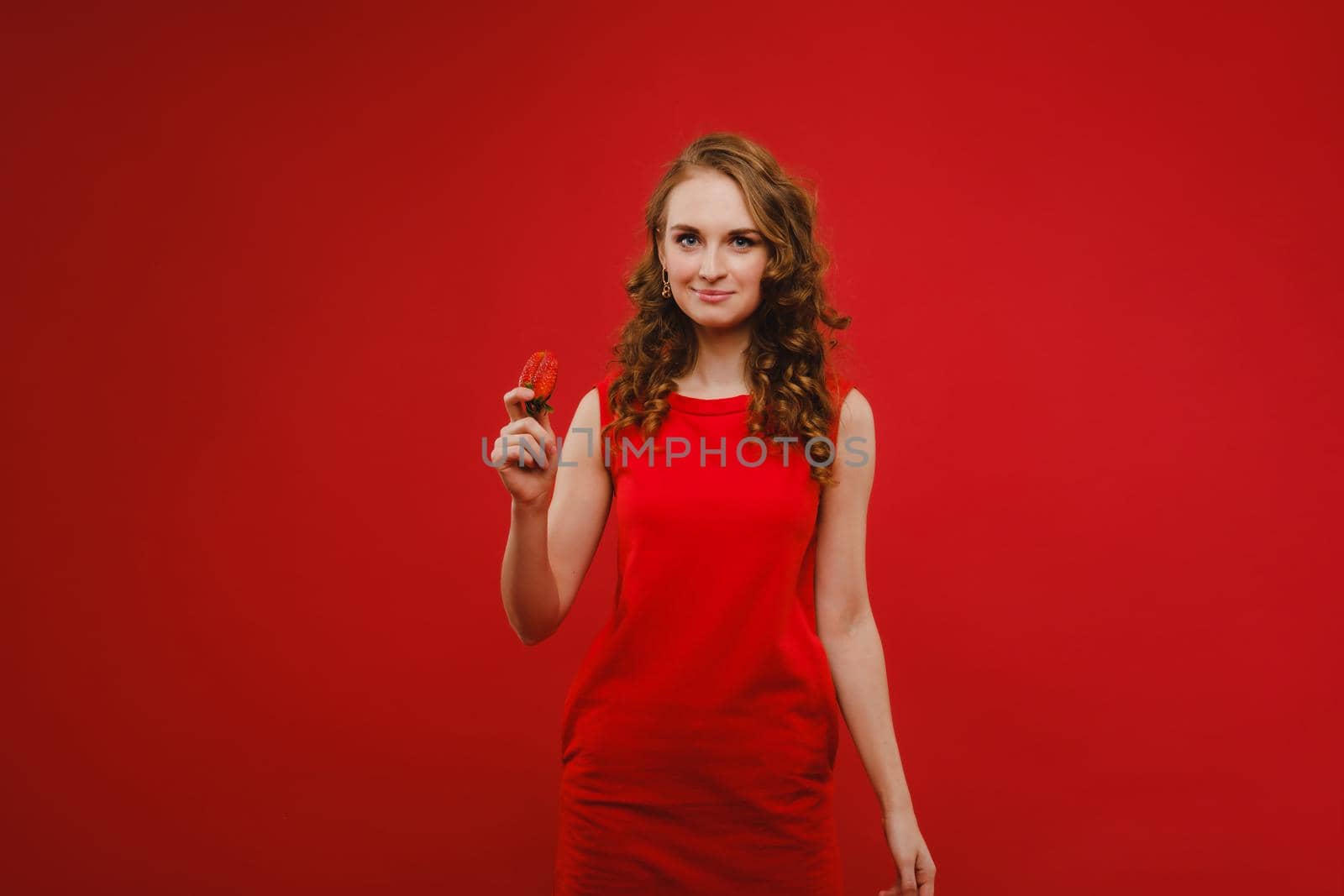 A beautiful girl in a red dress on a red background holds a strawberry in her hands and smiles.