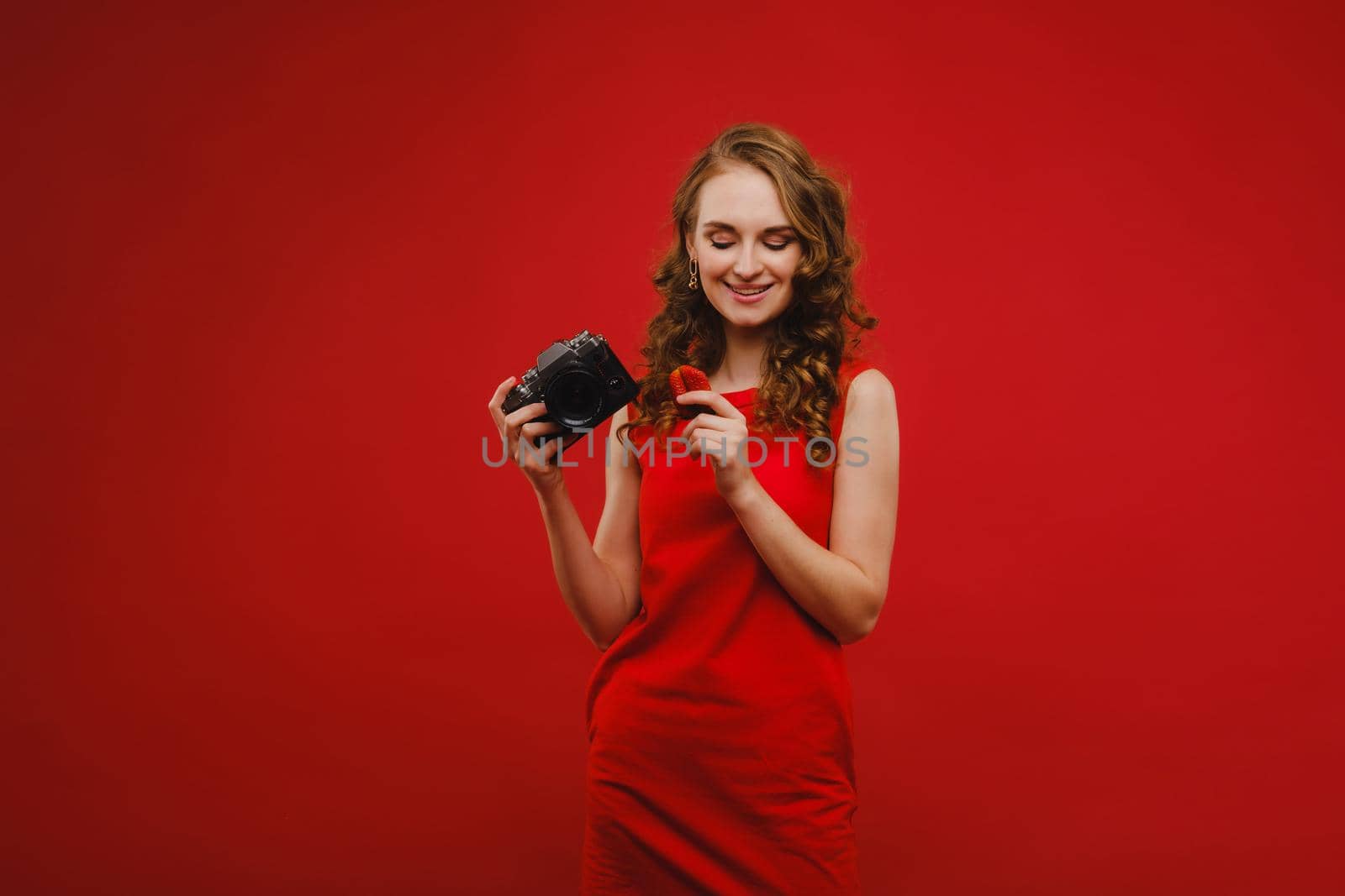 a smiling young woman with wavy hair holds a strawberry and photographs it, holding a delicious fresh strawberry on a bright red background.