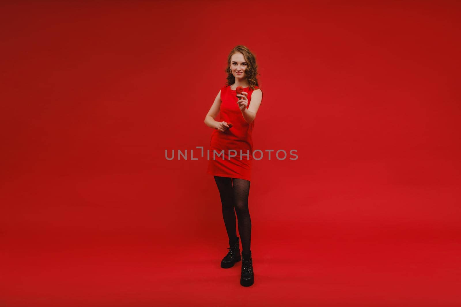 A beautiful girl in a red dress on a red background holds a strawberry in her hands and smiles.