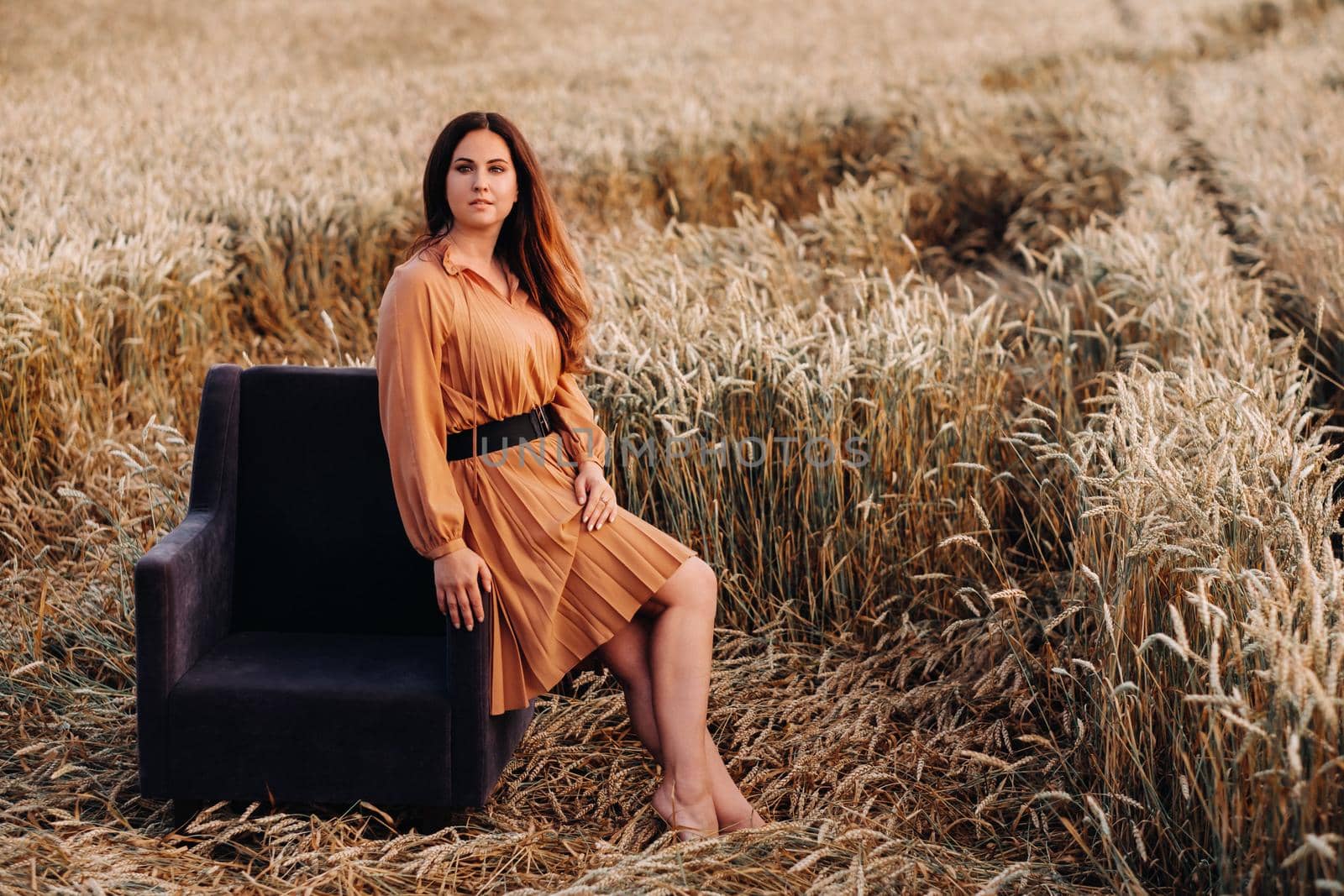 A girl in a orange dress is sitting on a chair in a wheat field in the evening.