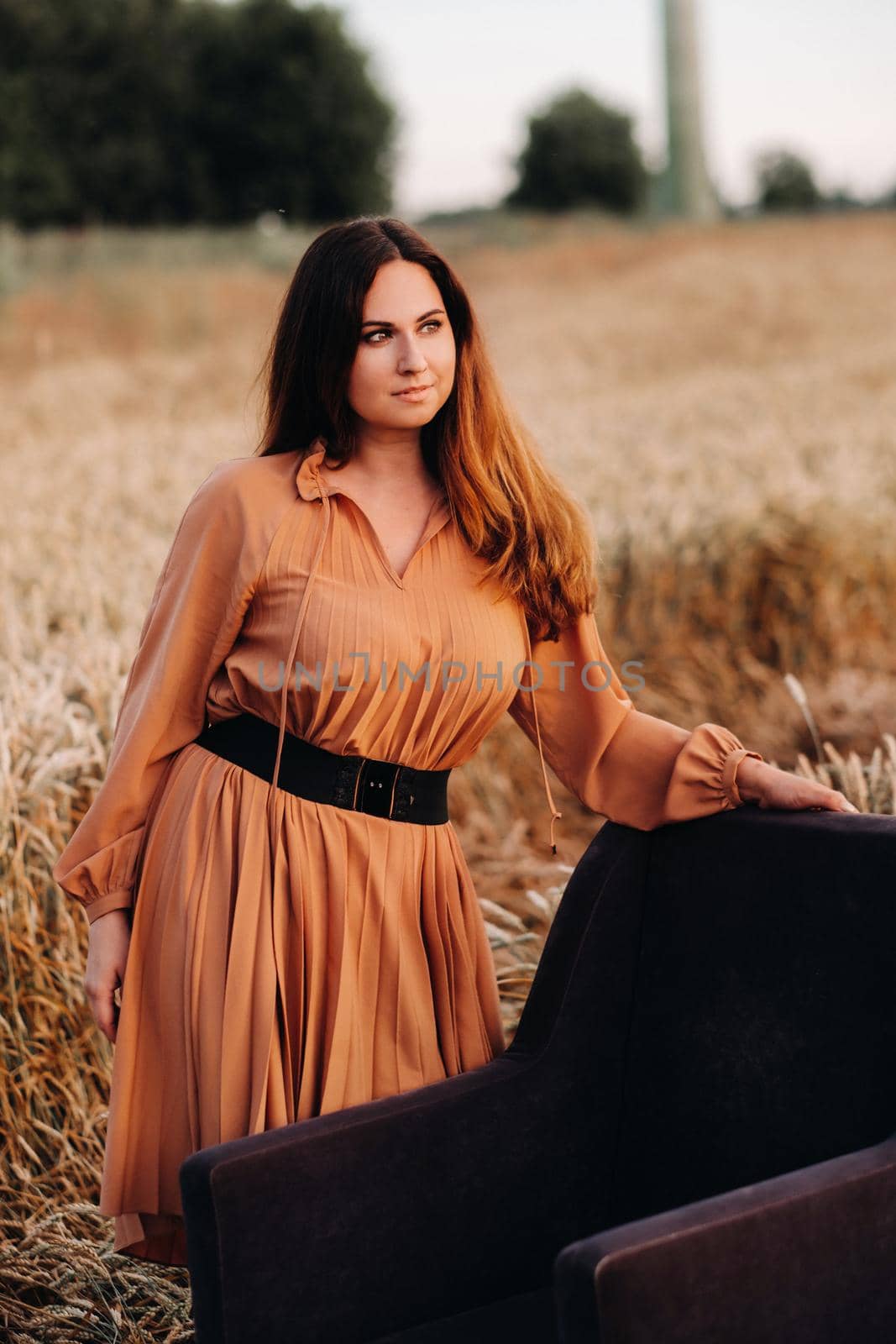 A girl in a orange dress stands next to a chair in a wheat field in the evening by Lobachad