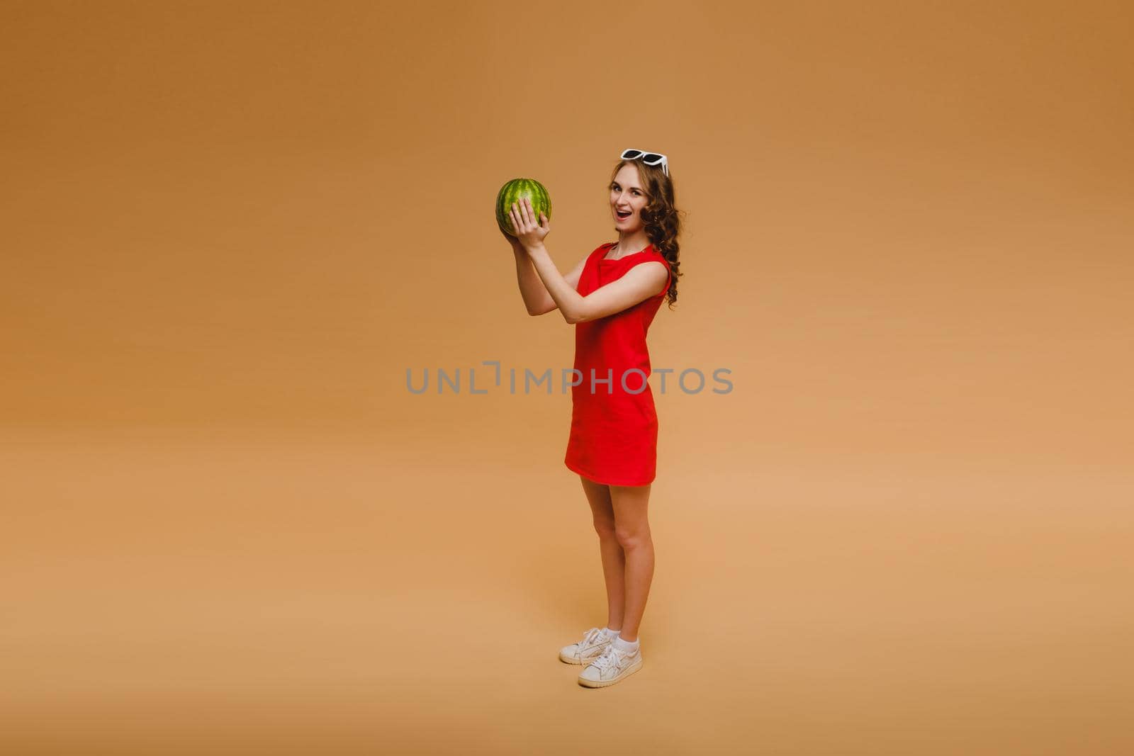 A beautiful girl in glasses and a red dress holds a watermelon in her hands.