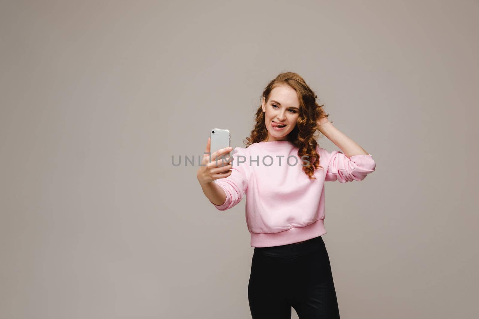 A smiling happy girl in a pink blouse takes a selfie on a smartphone on a gray background. by Lobachad