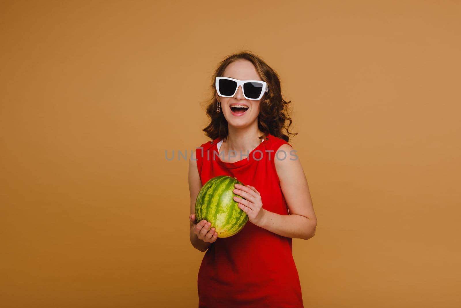 A beautiful girl in glasses and a red dress holds a watermelon in her hands.