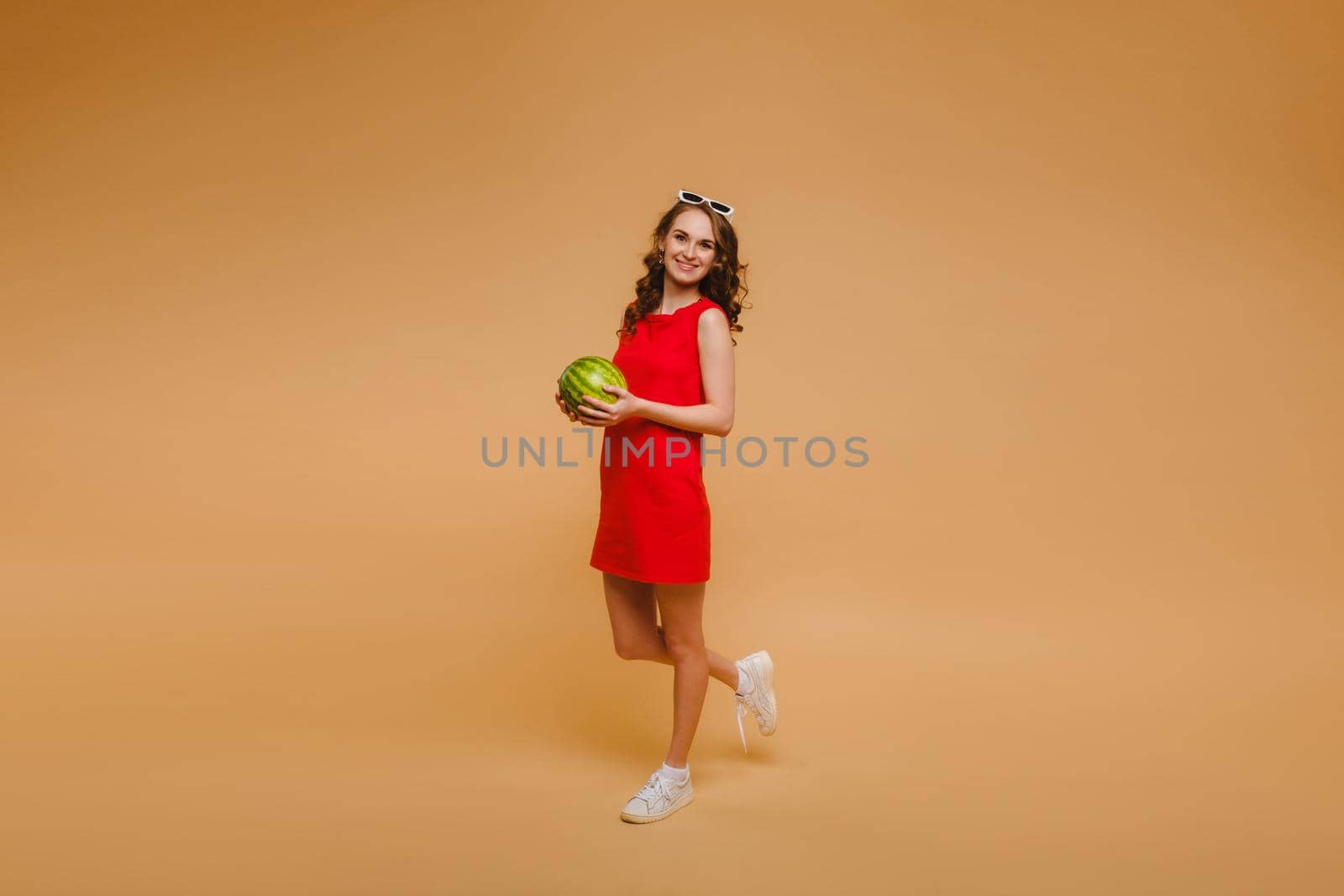 A beautiful girl in glasses and a red dress holds a watermelon in her hands.