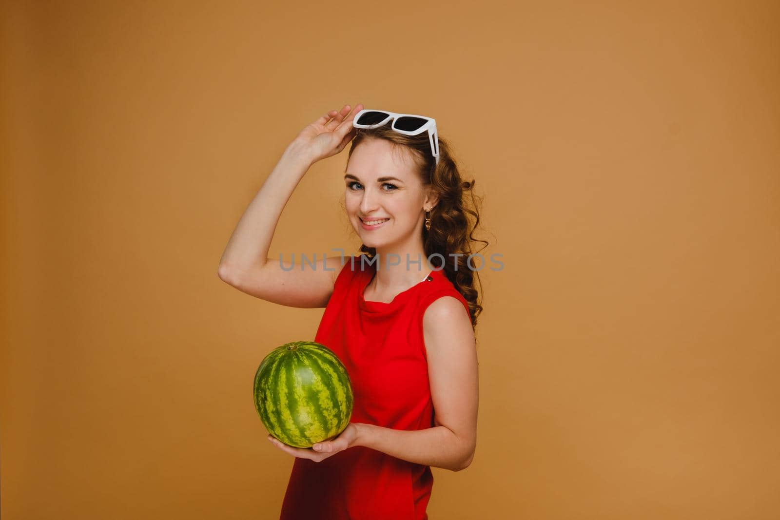 A beautiful girl in glasses and a red dress holding a watermelon on an orange background.