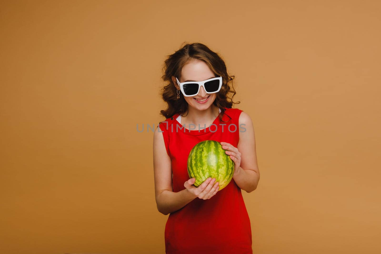 A beautiful girl in glasses and a red dress holds a watermelon in her hands.