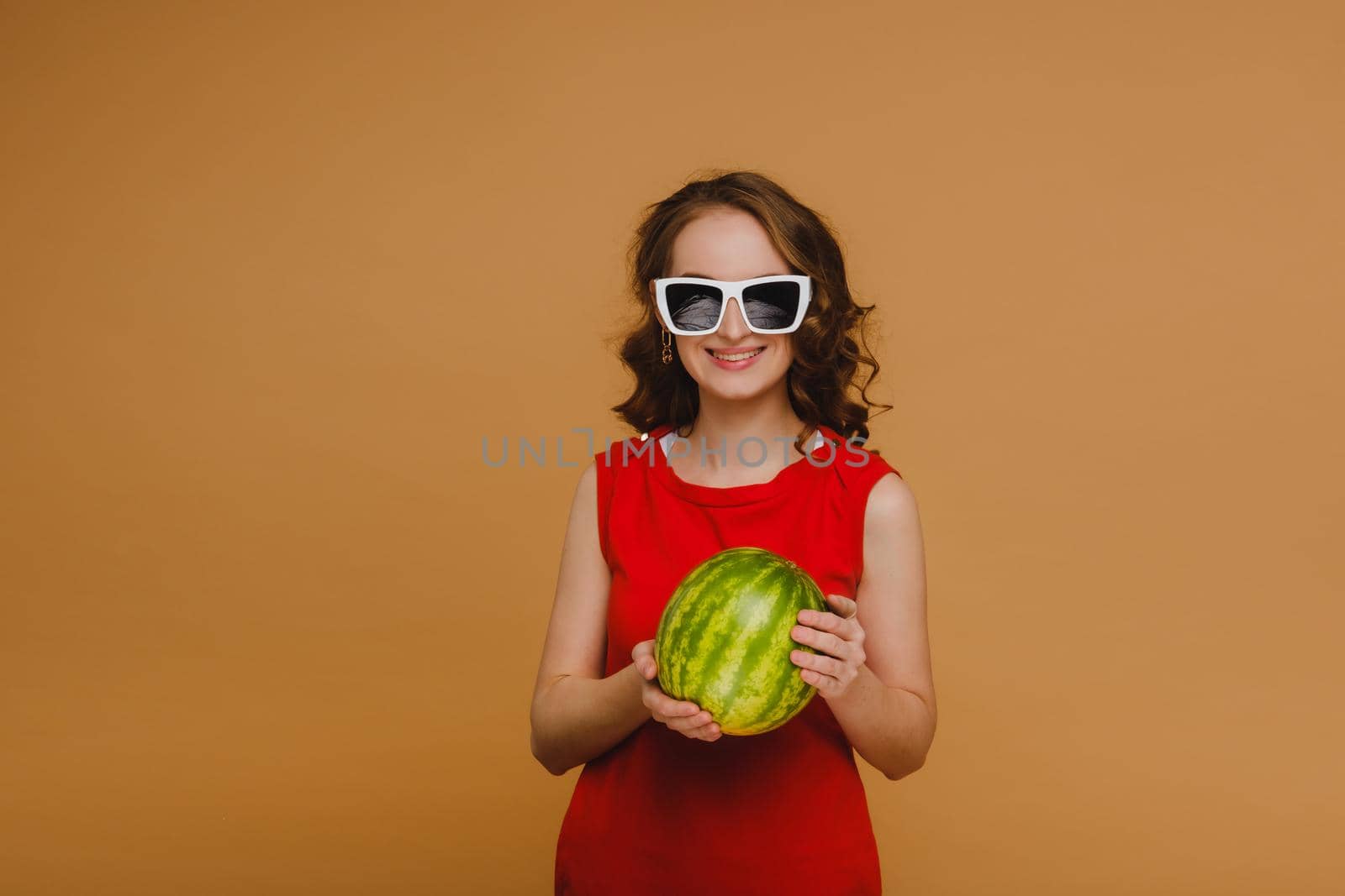 A beautiful girl in glasses and a red dress holds a watermelon in her hands.