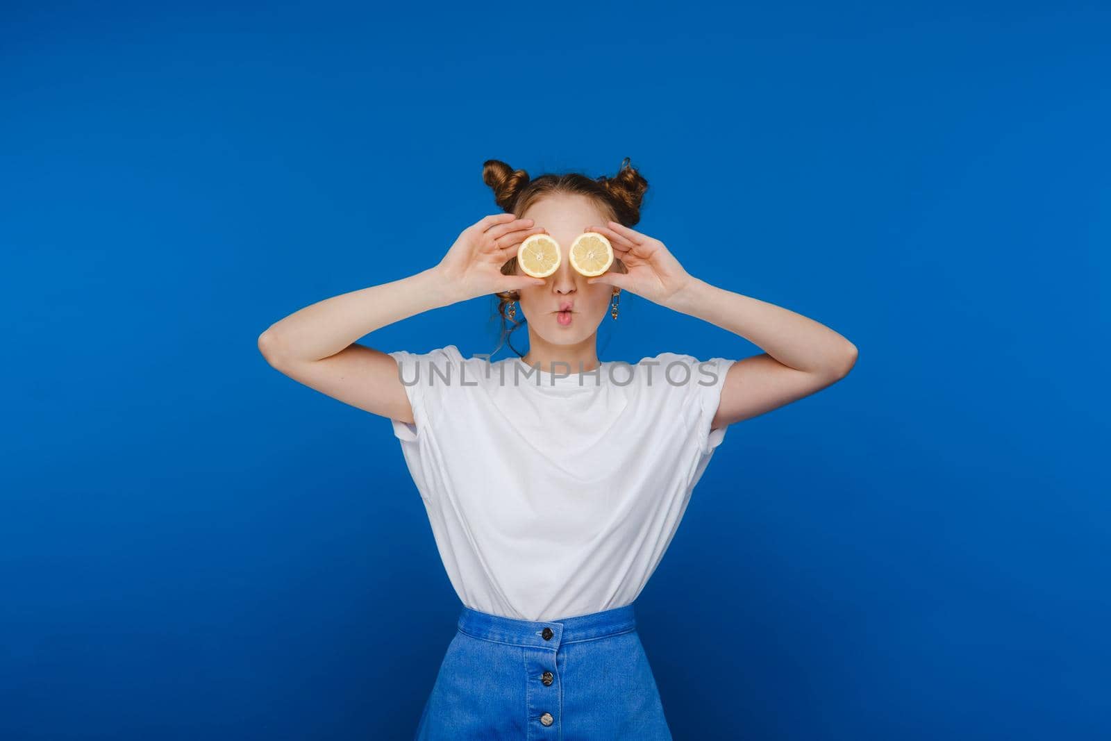 a young beautiful girl standing on a blue background holds lemons in her hands and covers her eyes with them.