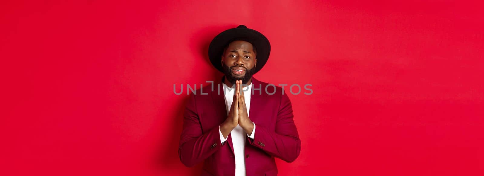 Miserable Black man asking for help, begging for favour and looking sad, standing against red background by Benzoix