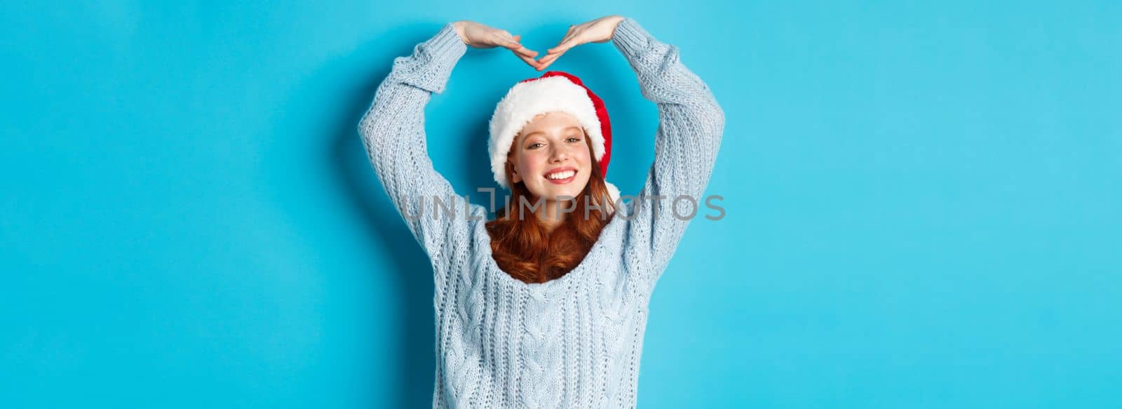 Winter holidays and Christmas Eve concept. Cute redhead teen girl in santa hat and sweater, making heart sign and smiling, wishing merry xmas, standing over blue background.