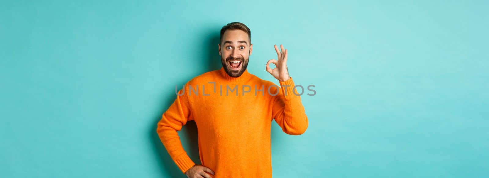 Impressed man recommending promo, showing okay sign and smiling amazed, approve and agree, standing over light blue background.
