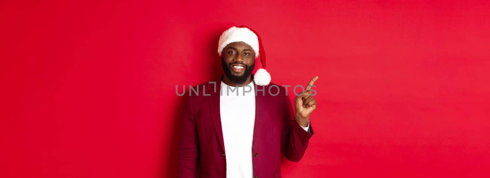 Christmas, party and holidays concept. Handsome Black man in santa hat pointing finger left, showing advertisement, standing happy over red background.