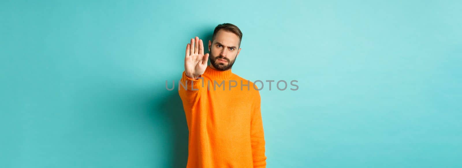 Serious frowning man showing stop gesture, telling no, prohibit and disagree, standing over light blue background.