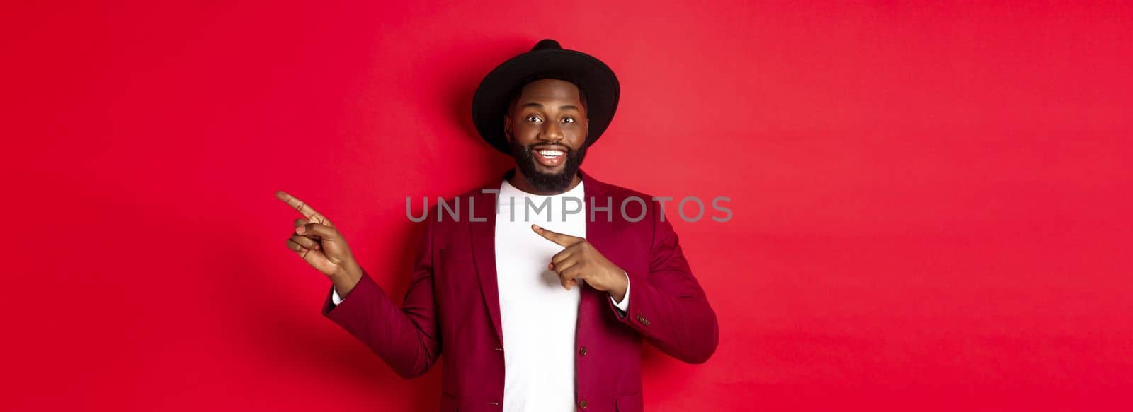 Winter holidays and shopping concept. Stylish african american man pointing fingers right at copy space for logo, standing over red background.