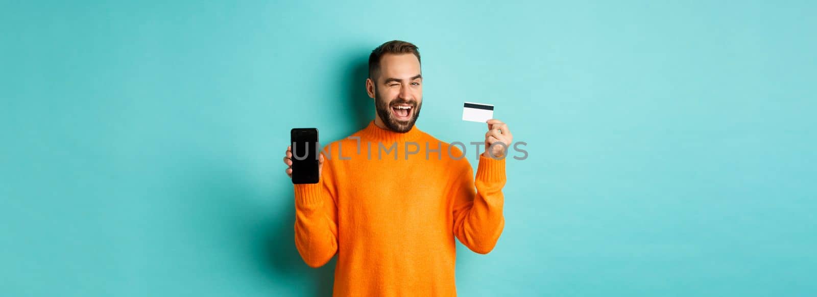 Online shopping. Handsome caucasian man showing credit card and smartphone, winking at camera, recommending something, standing over light blue background.