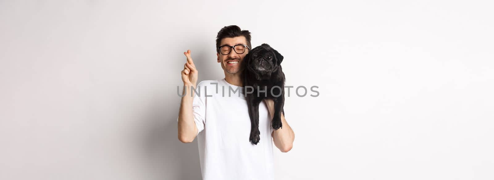 Hopeful smiling dog owner making a wish, holding cute black pug on shoulder and cross fingers for good luck, white background.