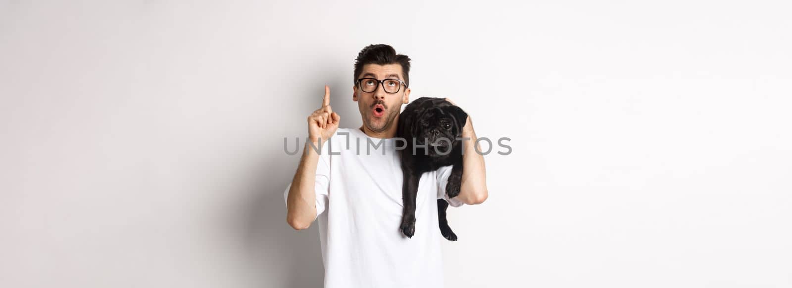 Amazed handsome man in glasses, holding cute black pug dog on shoulder, pointing finger up at promo logo, standing over white background.