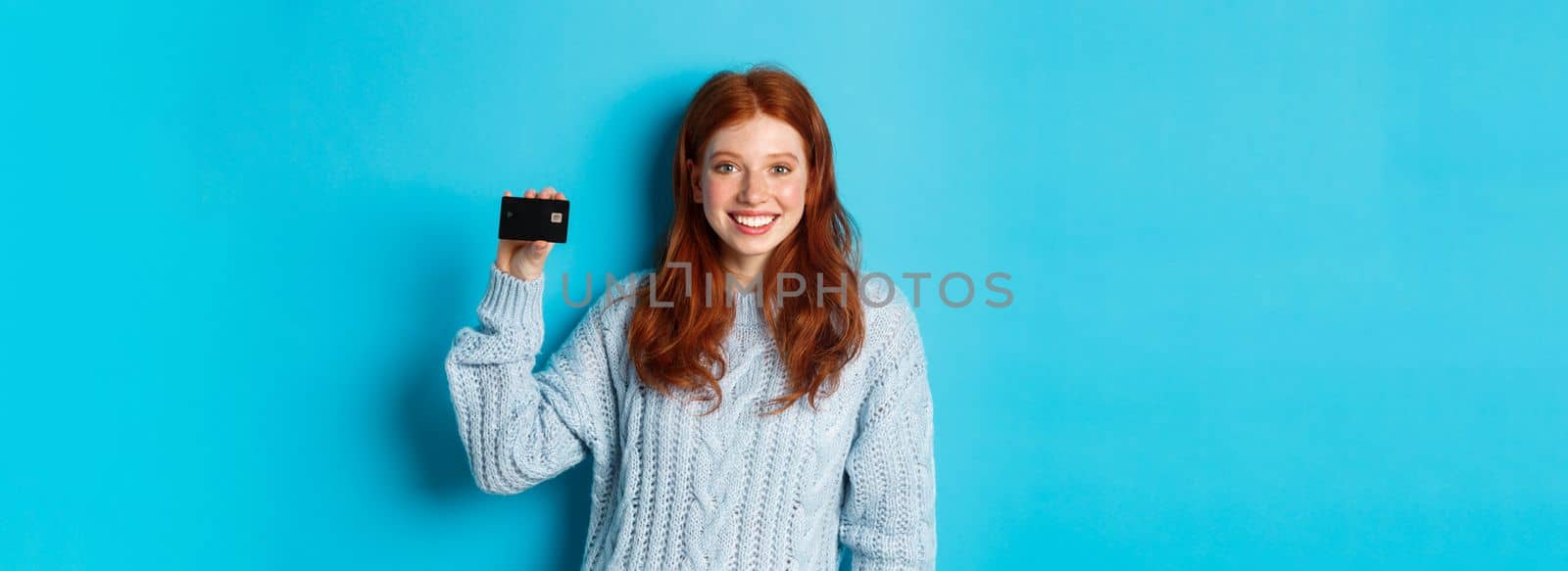 Cute redhead girl in sweater showing credit card, smiling at camera, standing over blue background.