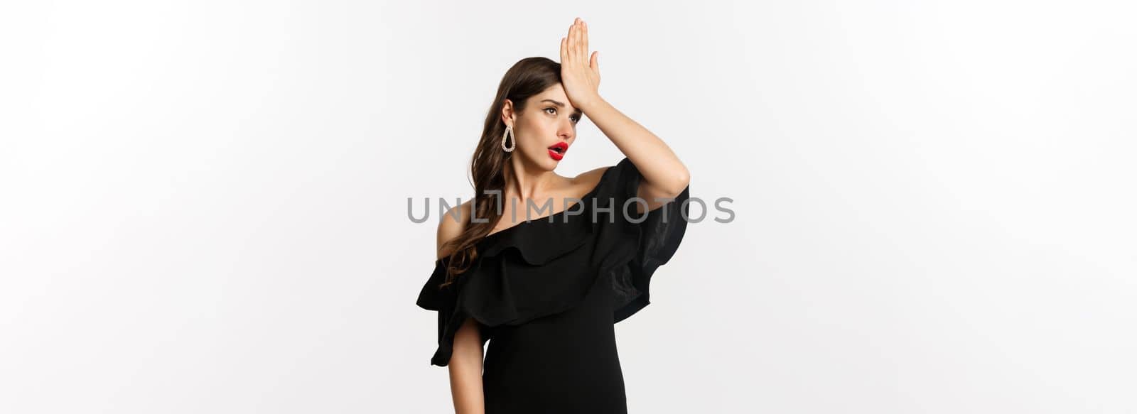 Fashion and beauty. Annoyed stylish woman in black dress, slap forehead, making facepalm and roll eyes tired, standing over white background.
