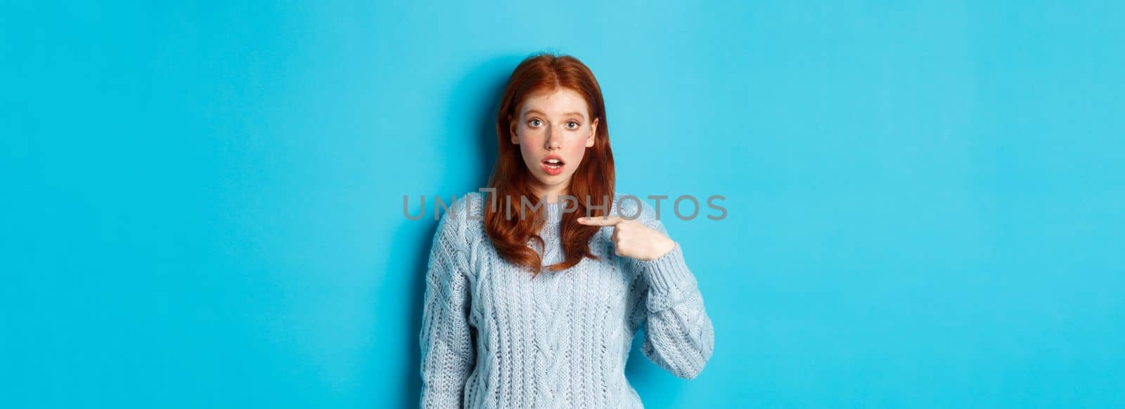 Confused redhead girl pointing at herself, being chosen, standing in sweater against blue background.