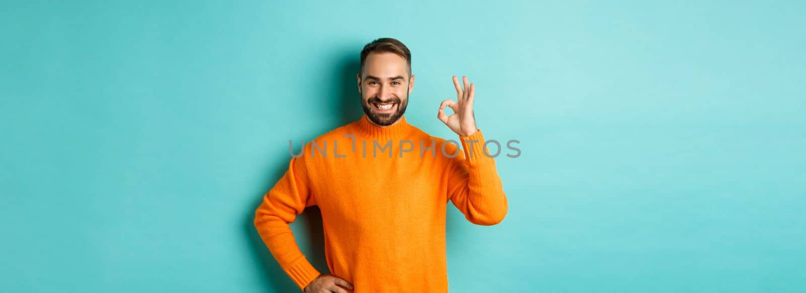 Image of confident handsome man showing ok sign, approve and agree, like something good, standing in orange sweater.