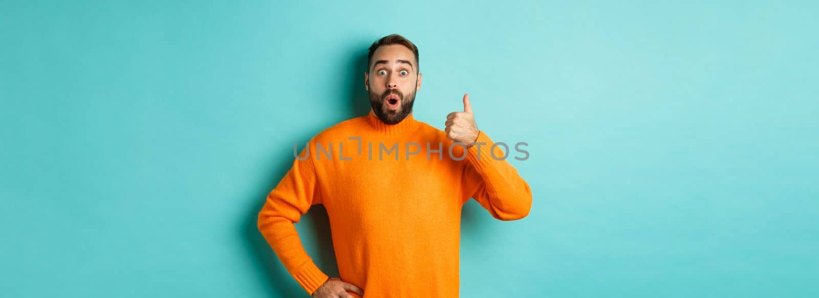 Well done. Handsome bearded man showing thumb up, praising good work, recommend excellent product, like and approve, smiling satisfied, standing over light blue background.