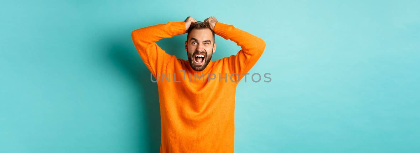 Frustrated man shouting, pulling out hair and screaming angry, losing temper and looking mad, standing over light blue background.