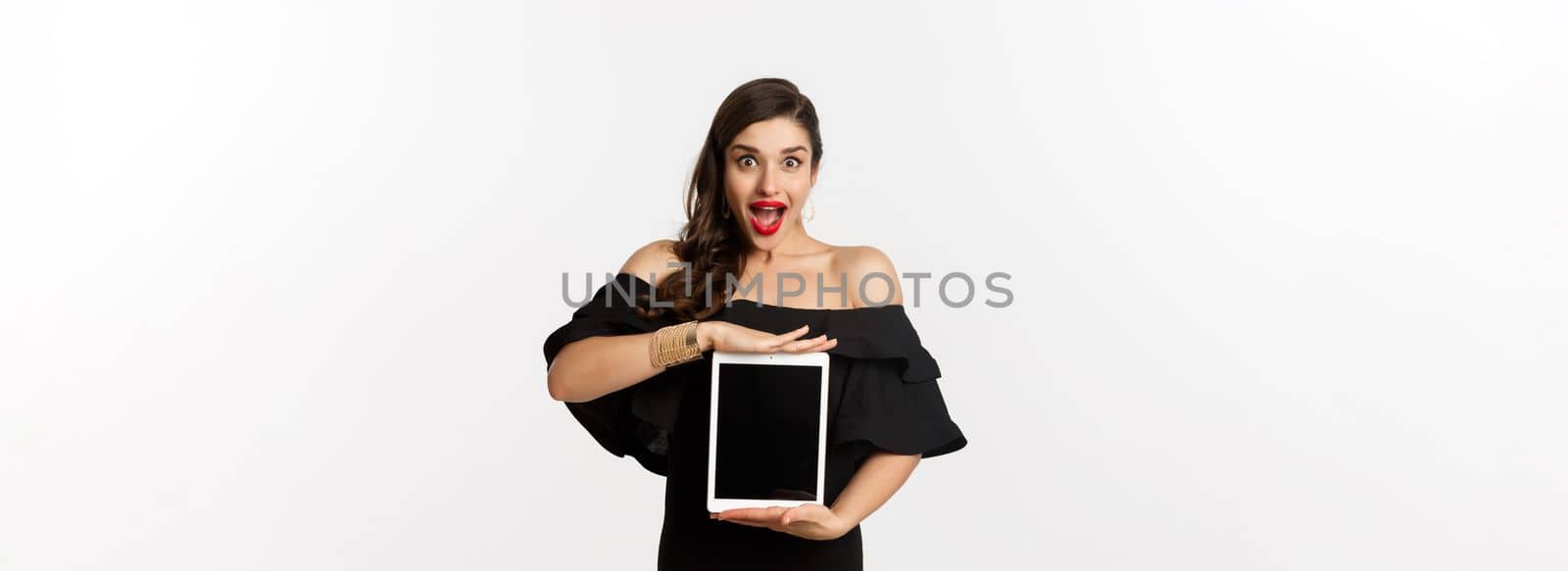 Fashion and shopping concept. Amazed young woman showing online website promo offer on tablet screen, looking excited camera, standing in black dress, white background.